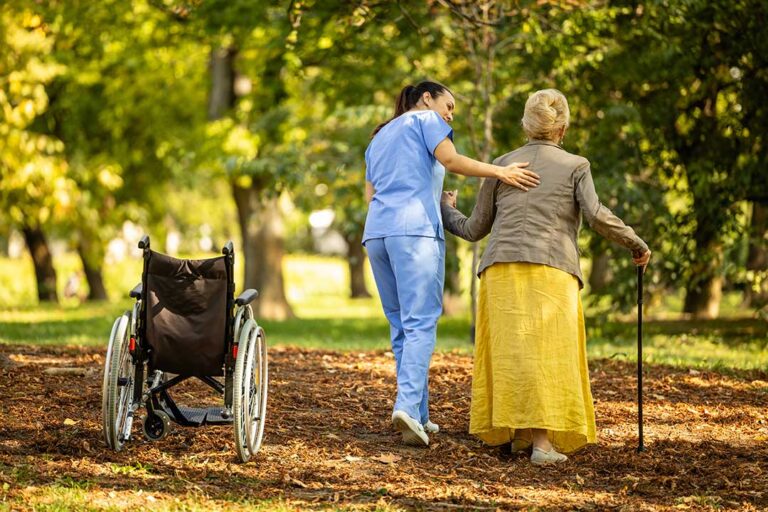 Caregiver calmly helps an older resident choose clothing in a bright assisted living room.